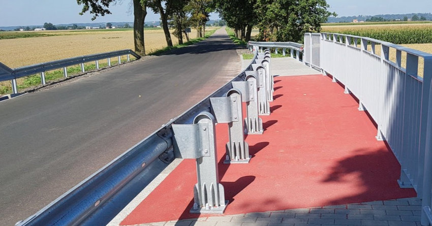 Fragment mostu z balustradą, obok droga asfaltowa ogrodzona z dwóch stron balustradą, w tle drzewa i pola.