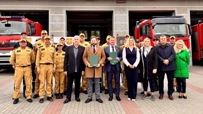 Na zdjęciu widać grupę ludzi pozujących do zdjęcia, na tle remizy strażackiej i wozów strażackich. W grupie są również umundurowani strażacy