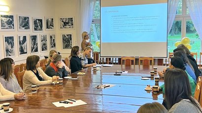 Na zdjęciu widać grupę uczestników szkolenia, siedzących przy dużym drewnianym stole, wpatrzonych w prezentację i prezenterkę. Na ścianie widać 11 obrazów przedstawiających podopiecznych warsztatów w Górkach.