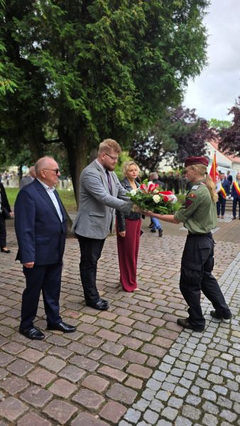 Zdjęcie przedstawia delegację 3 osób (2 mężczyzn i kobieta) przekazującą wiązankę kwiatów harcerzowi 