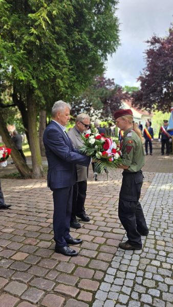 Zdjęcie przedstawia delegację 2 mężczyzn przekazujących wiązankę kwiatów harcerzowi 