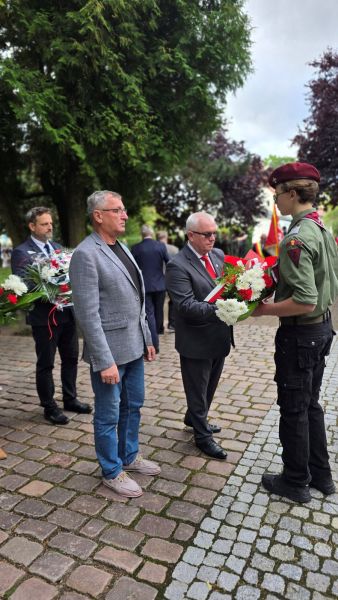 Zdjęcie przedstawia delegację 2 mężczyzn przekazujących wiązankę kwiatów harcerzowi 