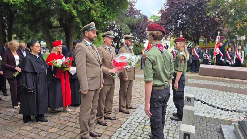 Zdjęcie przedstawia delegację 3 przedstawicieli nadleśnictwa przekazujących wiązankę kwiatów harcerzowi, za nimi delegacja 3 kobiet w togach