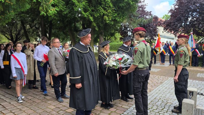 Zdjęcie przedstawia delegację 2 mężczyzn i kobieta, w togach przekazujących wiązankę kwiatów harcerzowi, za nimi widać kolejne delegacje z kwiatami