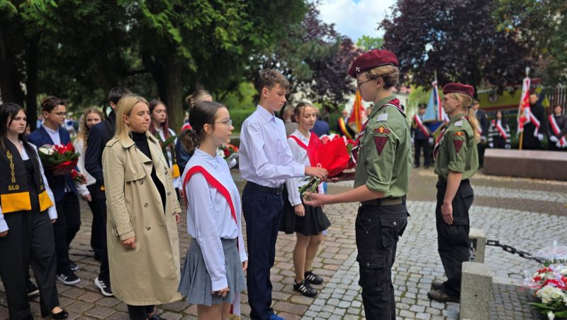 Zdjęcie przedstawia delegację uczniów, ubranych na galowo, przekazujących wiązankę kwiatów harcerzowi, za nimi widać kolejne delegacje