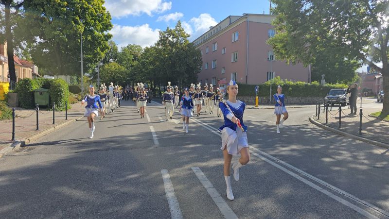 Zdjęcie przedstawia maszerującą ulicą orkiestrę dętą, na pierwszym planie 4 mażoretki