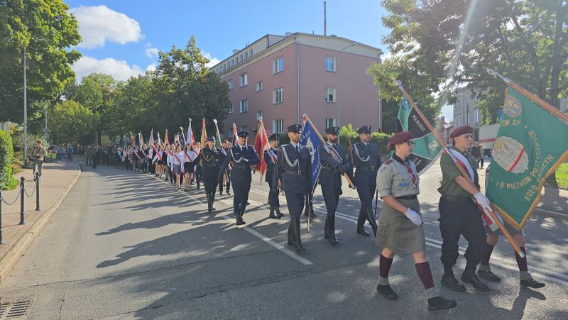 Zdjęcie przedstawia maszerujące delegacje ze sztandarami, na pierwszym planie delegacja harcerzy, za nimi mundurowi, dalej delegacje szkół