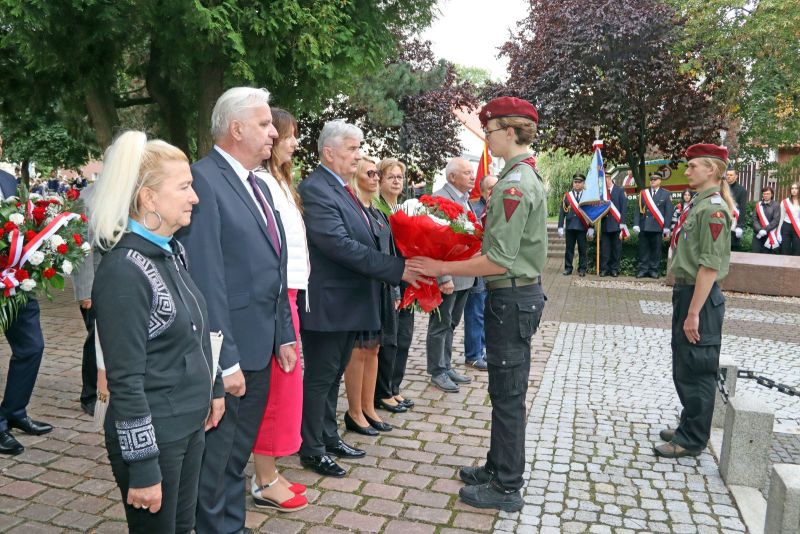 Zdjęcie przedstawia delegację 8 osób; Przewodniczący Rady Powiatu przekazuje harcerzowi wiązankę kwiatów pod pomnikiem