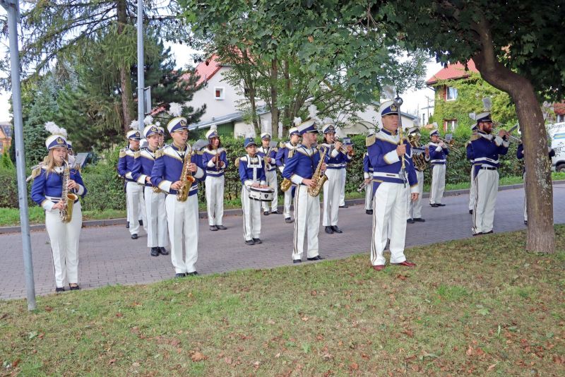 Zdjęcie przedstawia orkiestrę dętą stojącą na chodniku wśród drzew