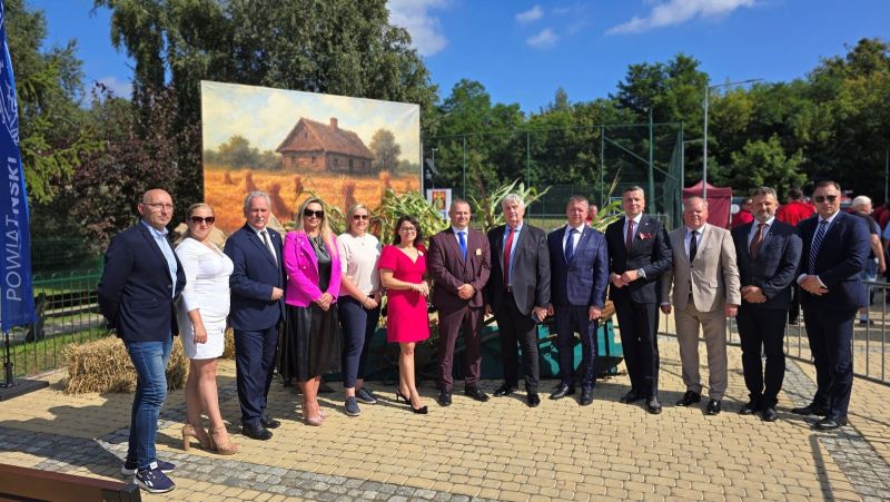 Grupa trzynastu osób, przedstawicieli Powiatu Kwidzyńskiego pozuje do zdjęcia na tle dekoracji dożynkowej.