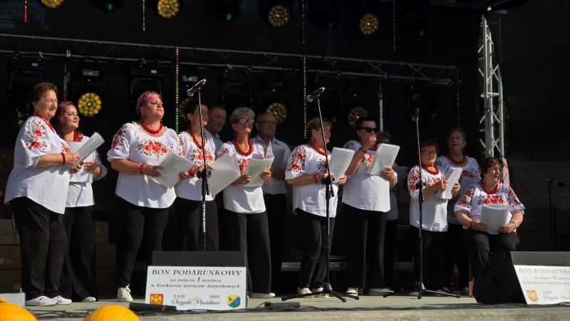 Zespół ludowy występuje na scenie, kobiety są ubrane w białe haftowane koszule, czarne spodnie i czerwone korale.