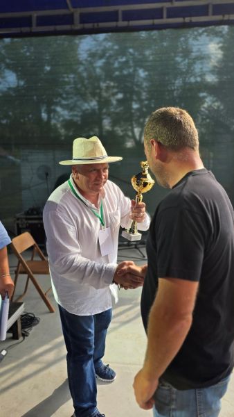 Mężczyzna w białej koszuli i słomianym kapeluszu wręcza puchar innemu mężczyźnie.