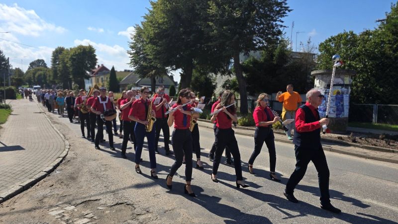 Przemarsz orkiestry dętej ulicą, członkowie zespołu ubrani w czerwone koszule i czarne krawaty.