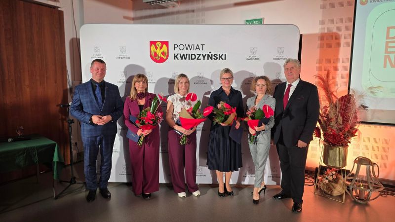 Na zdjęciu widać kobietę nagrodzoną z kwiatami w towarzystwie dwóch mężczyzn, w tle ścianka Powiatu Kwidzyńskiego