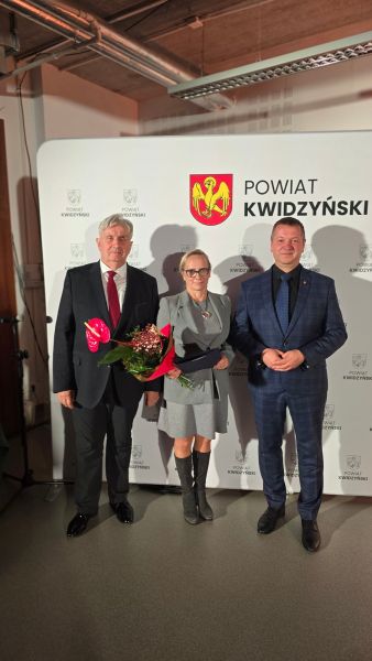 Na zdjęciu widać kobietę nagrodzoną z kwiatami w towarzystwie dwóch mężczyzn, w tle ścianka Powiatu Kwidzyńskiego