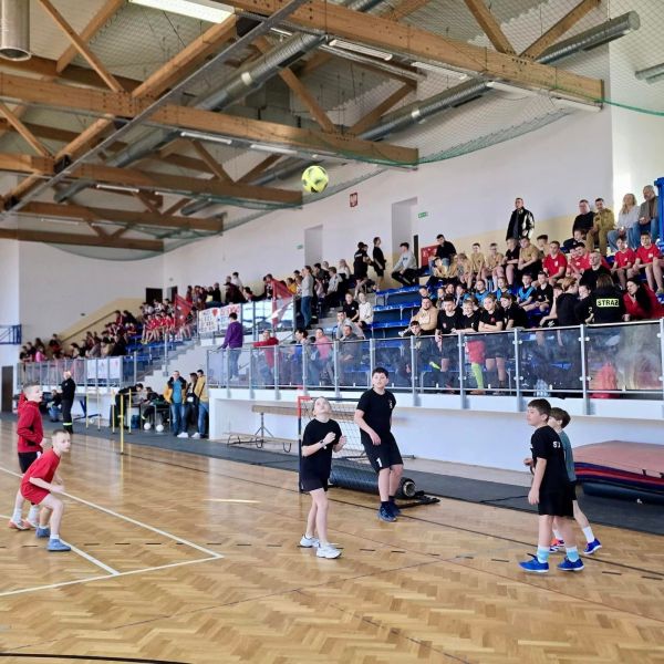 Zdjęcie przedstawia dzieci grające w piłkę nożną. W tle widzimy trybuny, a na nich wierni kibice drużyn. 