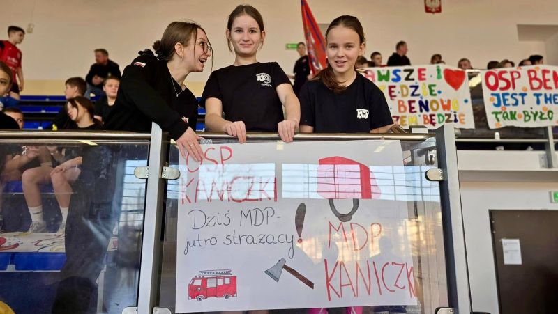 Zdjęcie przedstawia trzy młode dziewczyny, trzymające plakat przedstawiający radiowóz straży pożarnej, kask pożarniczy i siekierę. Na plakacie widnieje napis: OSP Kaniczki, Dziś MDP – jutro strażacy! MDP Kaniczki. 