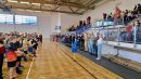 Zdjęcie przedstawia halę sportową, a po jej lewej stronie kolejno poustawiane drużyny sportowe. Po prawej stronie hali publiczność, trenerzy i organizatorzy wydarzenia. 
