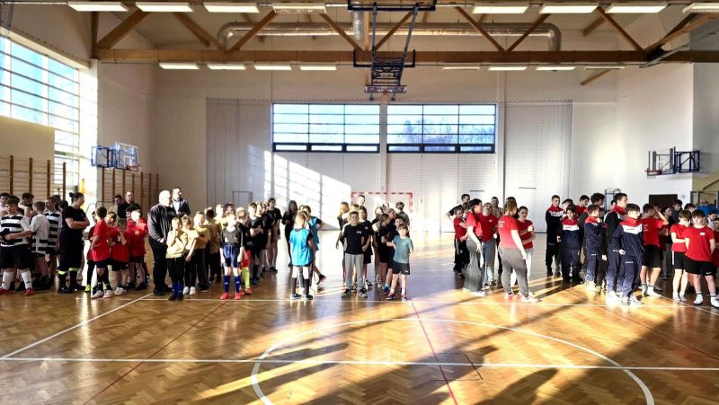 Zdjęcie przedstawia halę sportową i dzieci poustawiane w rzędach. Dzieci ubrane są w kolorach swoich drużyn. 