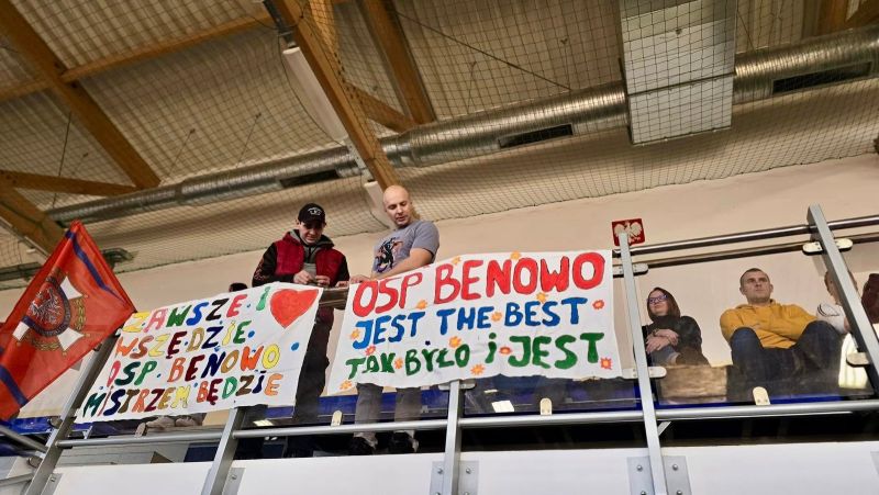 Zdjęcie przedstawia dwóch mężczyzn stojących na trybunach hali sportowej, a poniżej znajdują się dwa plakaty z napisem: Zawsze i wszędzie OSP Benowo Mistrzem będzie oraz OSP Benowo jest the best, tak było i jest. 