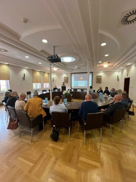 Zdjęcie przedstawia grupę siedzących osób przy okrągłym stole. W tle widnieje prezentacja. 