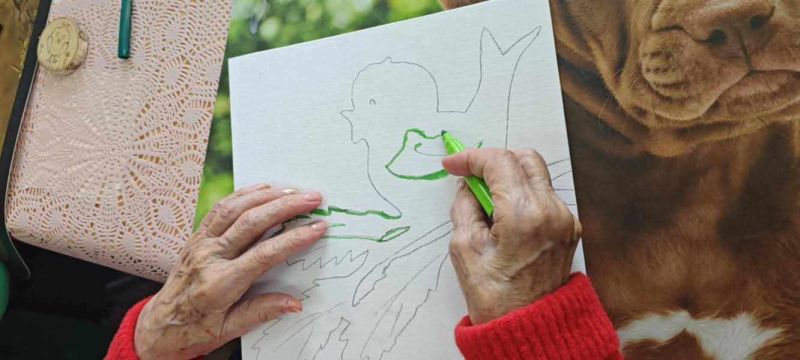 zdjęcie przedstawia dłonie starszej osoby, malującej pracę artystyczną. 