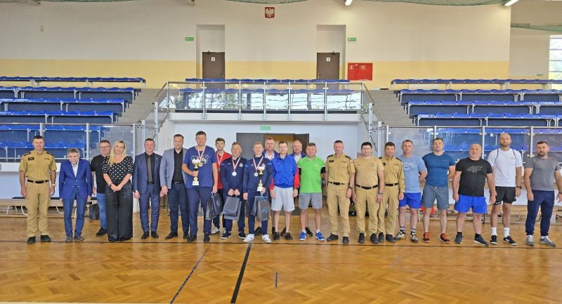 Grupa 22 osób pozuje do zdjęcia na hali sportowej, w tle widać trybuny, z przodu stoi trzech sportowców z medalami i torbami prezentowymi, dwóch z nich trzyma puchary.