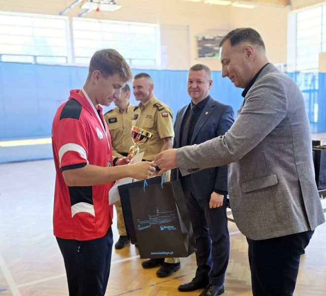 Burmistrz Miasta Kwidzyna wręcza mężczyźnie torbę upominkową, obok burmistrza stoi Starosta Powiatu Kwidzyńskiego, a dalej stoją dwaj strażacy.