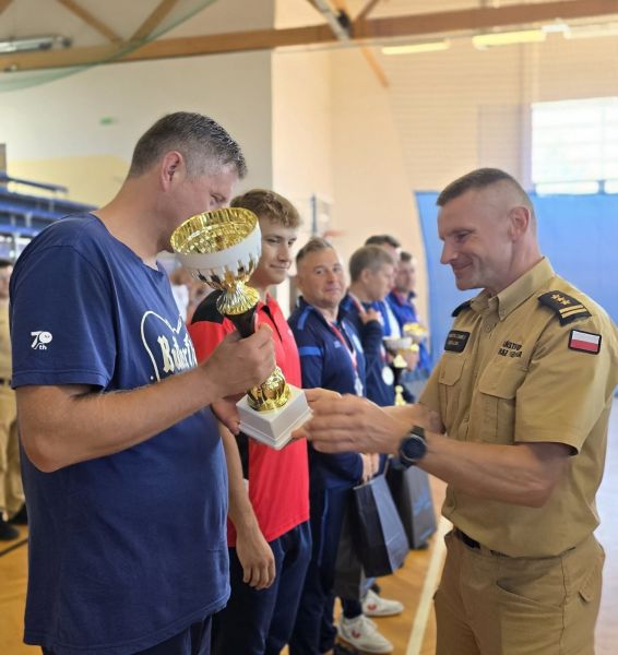 Przedstawiciel PSP w mundurze wręcza puchar sportowcowi, obok niego stoi w szeregu jeszcze 5 mężczyzn. 