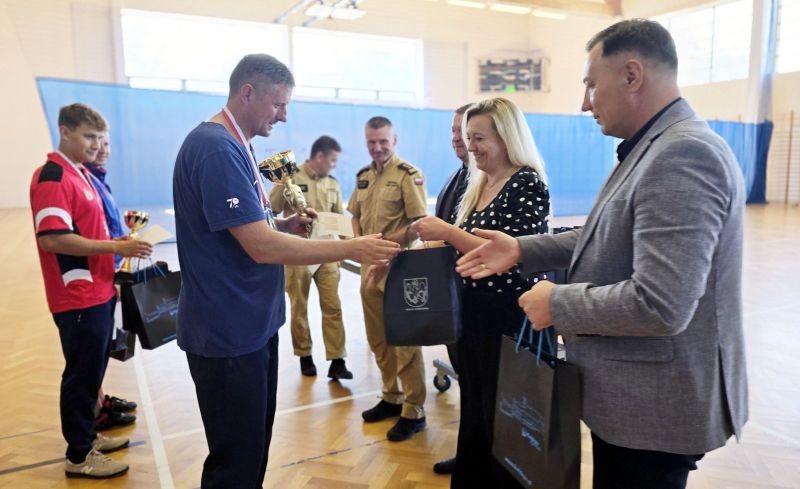 Członkini Zarządu Powiatu Kwidzyńskiego wręcza mężczyźnie torbę upominkową, obok burmistrz Miasta Kwidzyna wyciąga dłoń do gratulacji, w tle widać jeszcze pięciu mężczyzn.