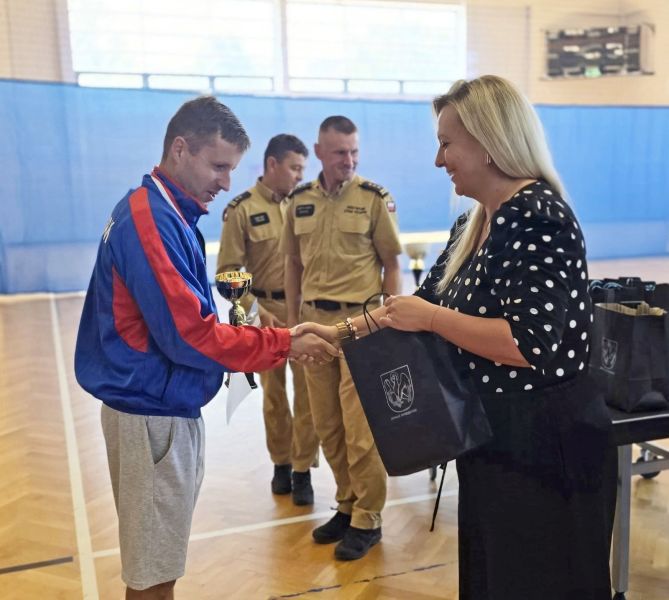 Członkini Zarządu Powiatu Kwidzyńskiego wręcza mężczyźnie torbę upominkową, w tle stoi dwóch strażaków.