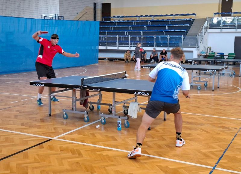 Dwóch mężczyzn gra w tenisa stołowego, w tle widać kobietę z rakietką przy drugim stole oraz trybyny.