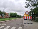 Znak „zakaz wjazdu” ustawiony przy modernizowanej drodze z widocznym zerwanym asfaltem. W tle widać budynki i drzewa.