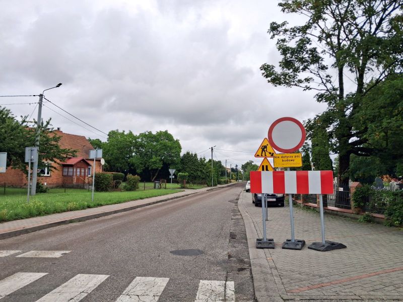 Znak „zakaz wjazdu” ustawiony przy modernizowanej drodze z widocznym zerwanym asfaltem. W tle widać budynki i drzewa.