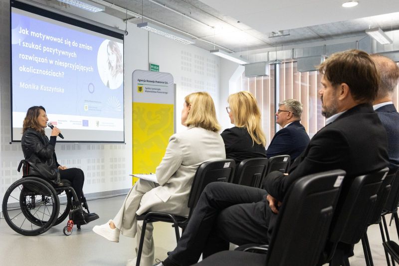 zdjęcie przedstawia osobę prowadzącą na wózku inwalidzkim i uczestników konferencji.