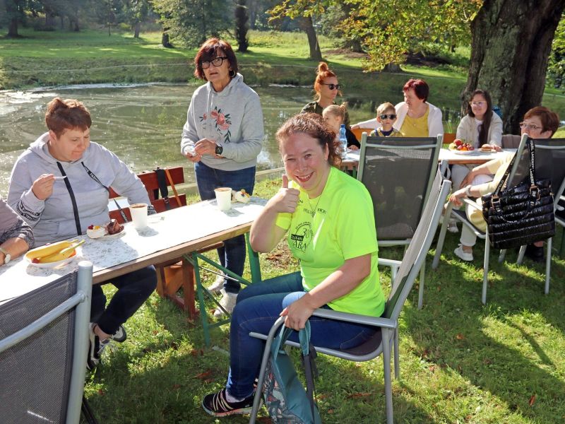 Zdjęcie przedstawia grupę ludzi siedzących na pikniku nad stawem, na pierwszym planie uśmiechnięta kobieta pokazująca kciuk skierowany do góry