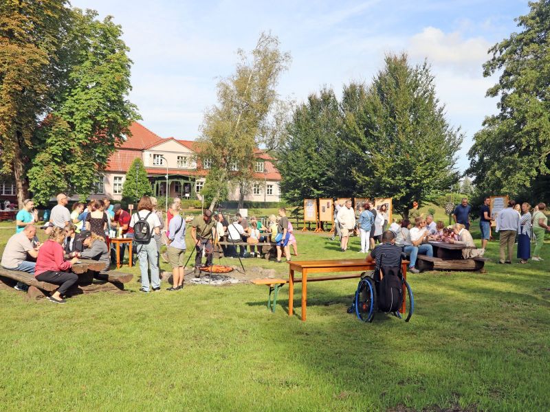 Zdjęcie przedstawia grupę ludzi podczas pikniku na dworze, z przodu widać mężczyznę na wózku siedzącego tyłem, część ludzi siedzi przy stołach; mężczyzna przekłada kiełbaski na ruszcie ogniska