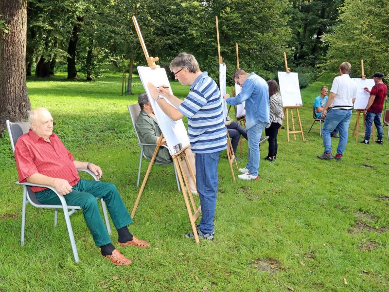 Zdjęcie przedstawia malarzy malujących na sztalugach oraz modeli siedzących na krzesłach, w tle piękna przyroda 