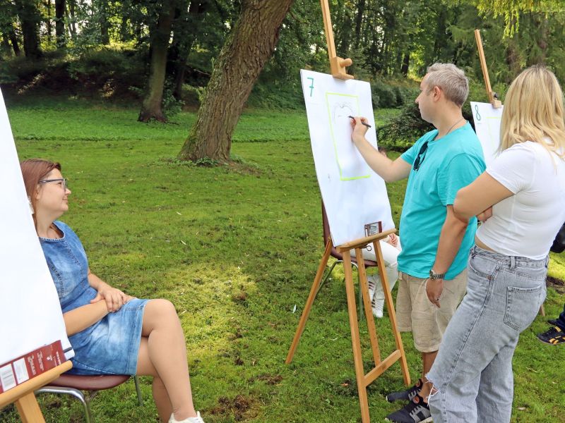 Zdjęcie przedstawia mężczyznę malującego na sztaludze w otoczeniu przyrody, obok na krześle siedzi modelka, za malarzem stoi odwrócona tyłem kobieta