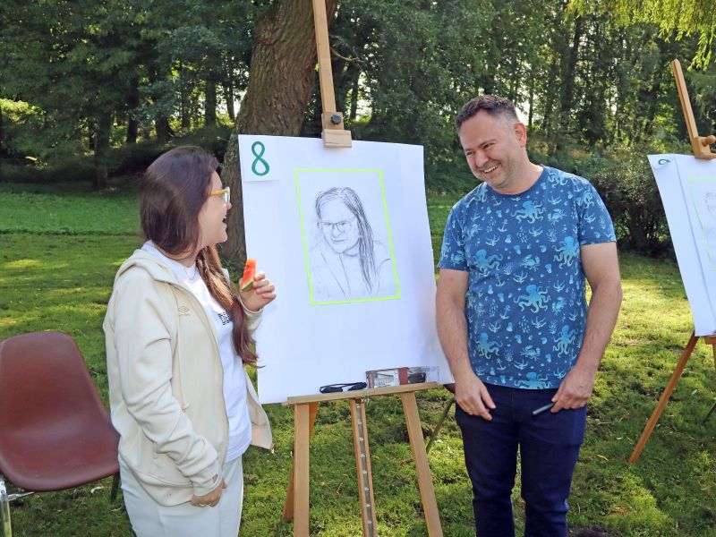 Zdjęcie przedstawia kobiety i mężczyzny stojących po dwóch stronach sztalugi, na rysunku portret stojącej obok kobiety