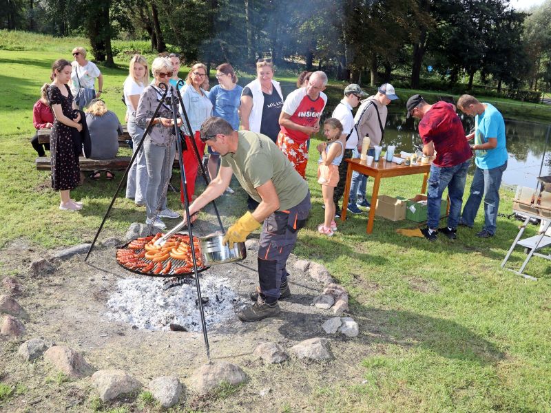Zdjęcie przedstawia mężczyznę, który przekłada na ruszcie kiełbaski, w tle grupa ludzi oraz staw