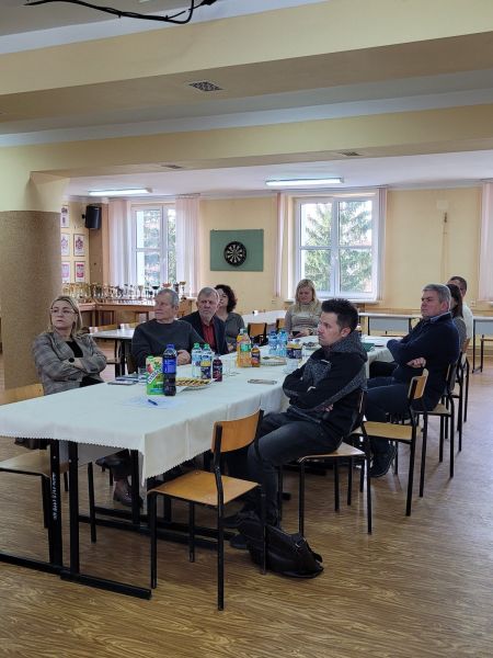 Grupa osób siedzi przy stole i słucha prezentacji w sali z dużymi oknami, pucharami i herbami na ścianach.