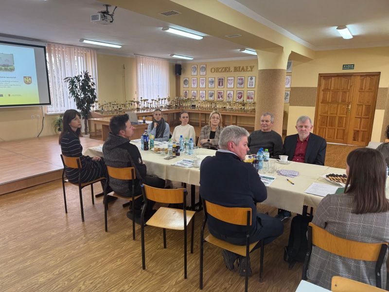 Uczestnicy spotkania nauczycieli siedzą przy stole i rozmawiają, w tle ekran z prezentacją i napis ‘Orzeł Biały’ na ścianie.