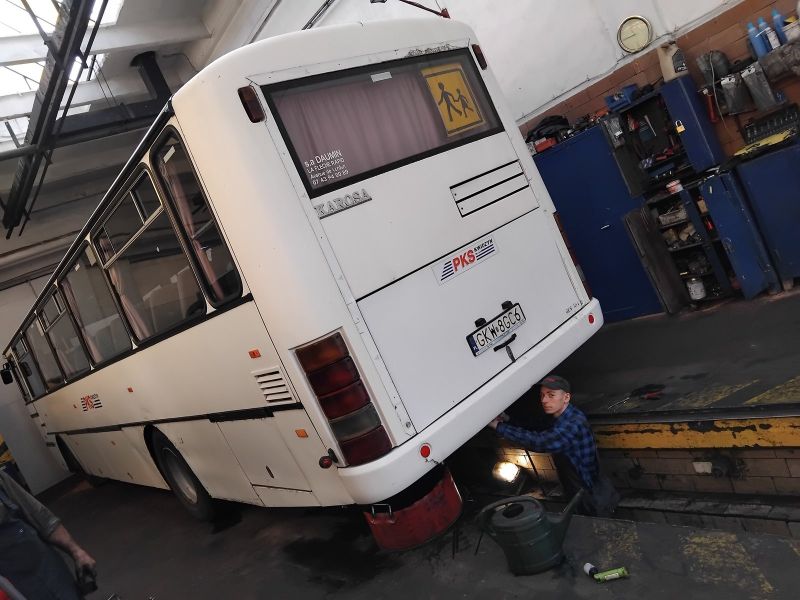  zdjęcie przedstawia stażystę podczas stażu uczniowskiego, znajduje się on w kanale w zakładzie samochodowym i sprawdza podwozie  autobusu;