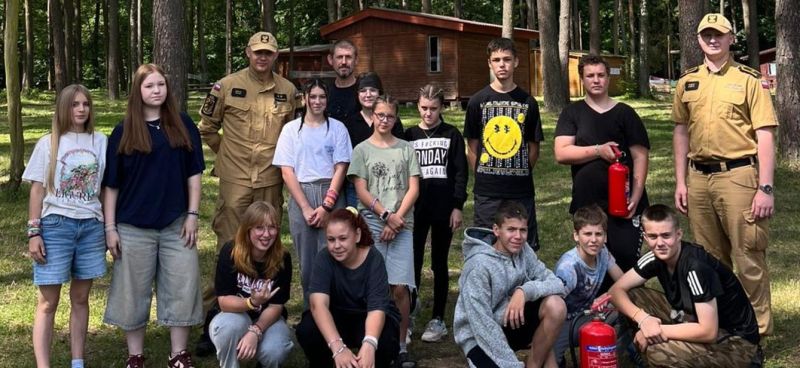 Harcerze i strażacy pozują do wspólnego zdjęcia pamiątkowego w obozowej scenerii.