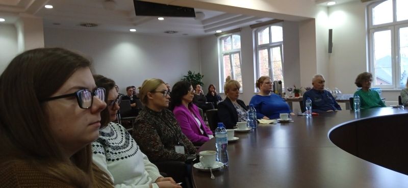 Druga grupa osób uczestnicząca w szkoleniu na miejscach siedzących przy okrągłym stole konferencyjnym oraz w tylnej części sali