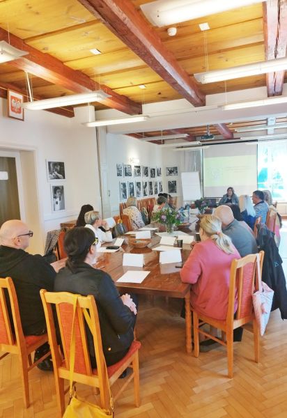 Uczestnicy siedzą przy jednym stole i słuchają prowadzącej szkolenie.